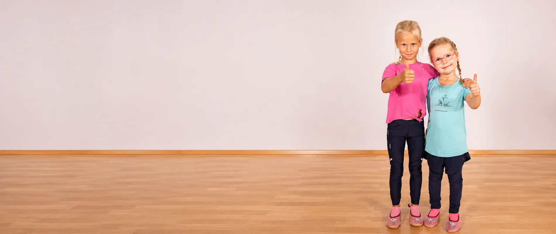Zwei fröhliche Mädchen beim Kindertanzkurs in der Tanzwelt Regensburg – mit Daumen hoch vor einer hellen Tanzstudio-Wand und Parkettboden.